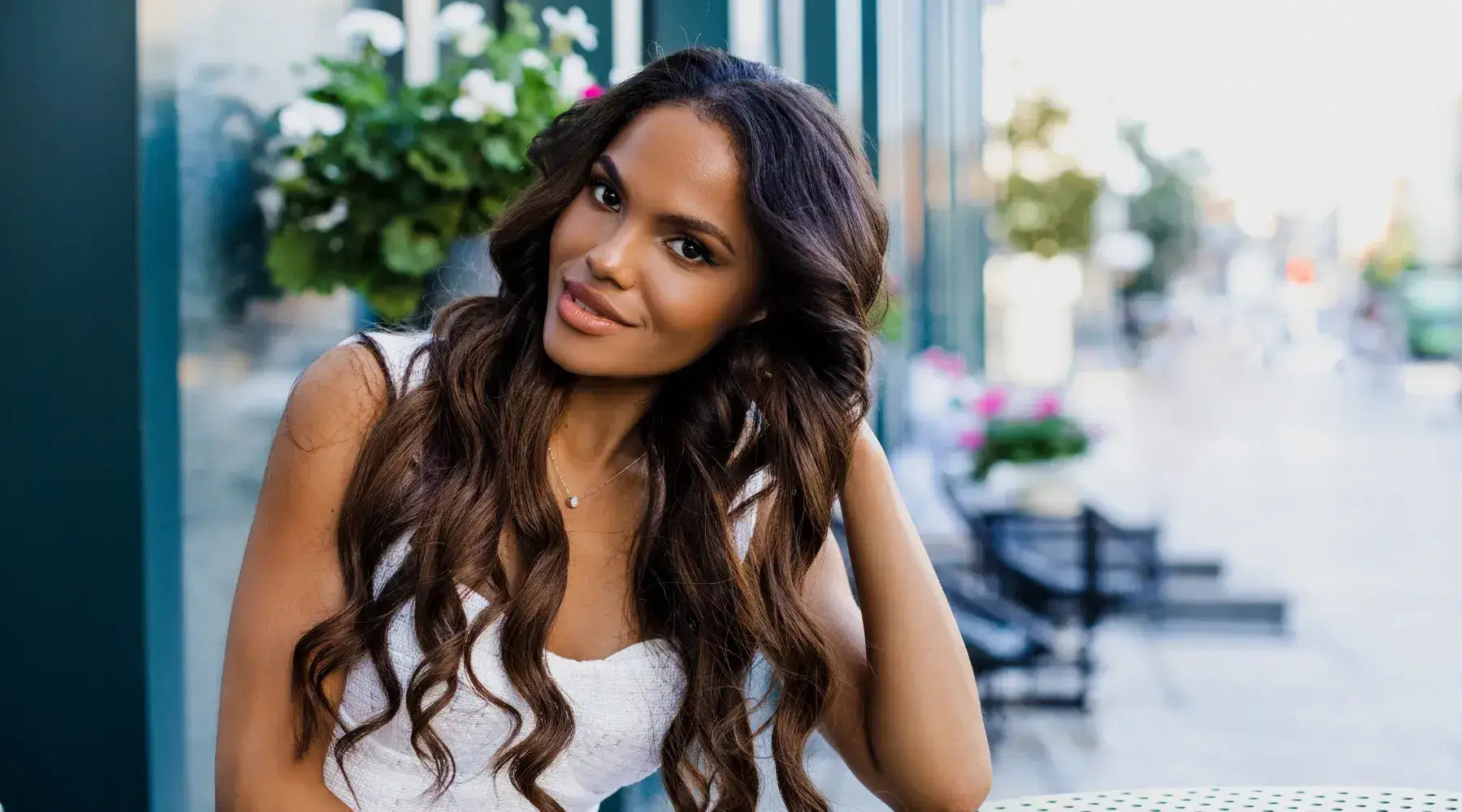 A stunning Black model with a long dark and wavy hairstyle sits at an outdoor table